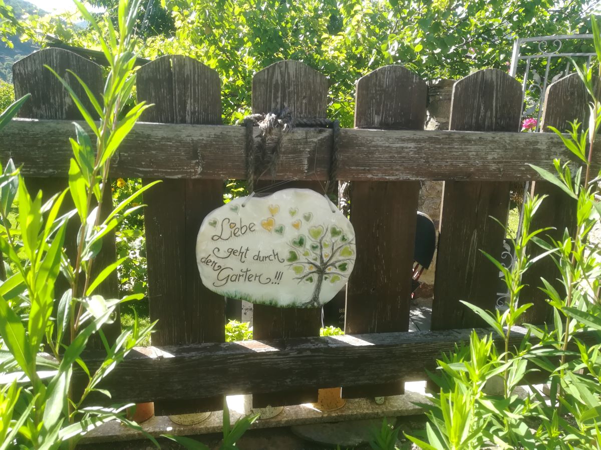 Keramikschild mit der Aufschrift "Liebe geht durch den Garten!" an einem Holzzaun.